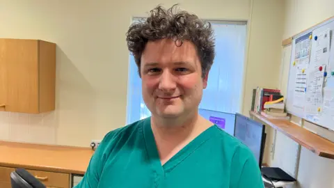 A GP with brown curly hair wearing blue overalls is standing inside a GP clinic and smiling at the camera. Behind him is a wooden table, office chair, computer and whiteboard with several pieces of paper attached to it. 