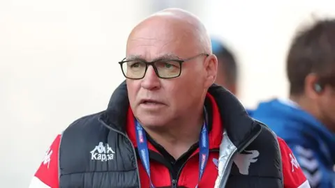 PA A man with square black glasses wearing a rugby jersey and gilet. He is bald and has a focused expression on his face.