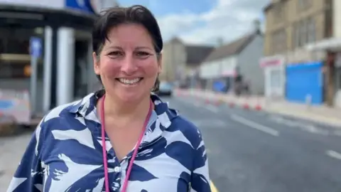Nicola Haseler/BBC Andrea Spice with medium-length dark hair, wearing a blue and white top and pink lanyard, smiling at the camera. A town street is visible behind her, with some road cones alongside the road.