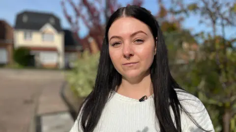 A woman with long dark hear, a nose ring and wearing white jumper. She is looking directly at the camera