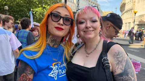 Eva, who has bright dyed orange hair, wears black sunglasses, red lipstick and a blue top as she smiles in a posed photo with Pippa, who has short pink dyed hair and is also smiling