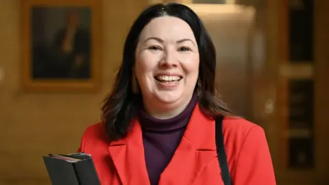 Getty Images Monica Lennon, who has shoulder-length dark hair, smiles while looking at the camera. She is wearing a red jacket and carrying a black folder 