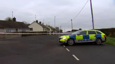 BBC A police Car sits infront of a group of white brick houses. Inbetween the car and the houses is red and white police tape. 