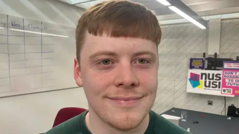 A man with ginger hair wearing a green t-shirt looks at the camera. There are NUS signs and a whiteboard behind him.