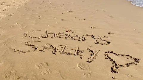 Rainbow Watcher A sandy beach with the words "Cheers Sally" and a love heart drawn. It is in relation to Sally Taylor hosting her final BBC South Today this week.