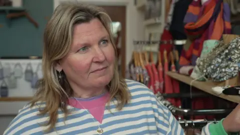 BBC/Luke Manterfield Rachael Cumming, who has blonde hair and is wearing a blue striped top, standing in her shop, there are clothes on rails behind her.