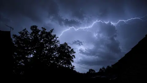 Freya N A bolt of lightning across a light-blue clouded sky during the night. Trees and houses can be seen on the ground.