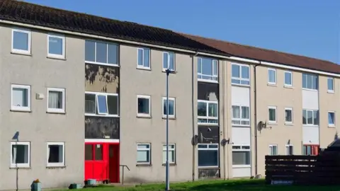 A block of flats in an urban setting. They are mostly grey with some white window columns and one has a red door. There is a patch of grass and a lamppost in front of the flats.