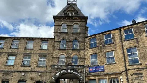 A large former mill building, made of light-coloured stone, with several floors and a large tower. Above the building arch, two signs read: Wellington Mills and Heritage Exchange. 