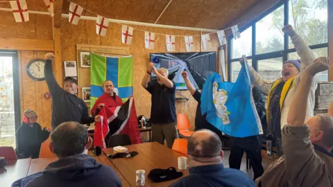 A group of people in a wood‑panelled room cheer and raise flags while watching a broadcast of the Winter Olympics on a wall‑mounted TV. 
