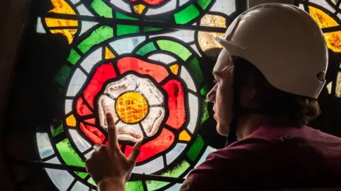 Danny Lawson/PA Wire Apprentice glazier Theo Dives, from York Glaziers Trust, wears a white hard hat. He points to a small section of the Rose Window's stained glass in the shape of a red, white and yellow rose
