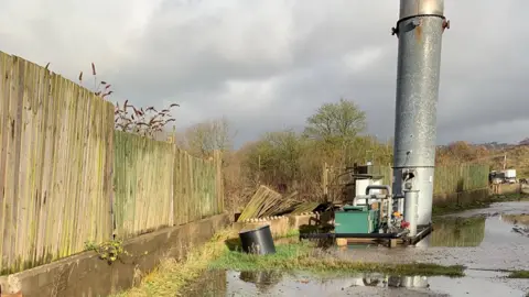 Environment Agency An outdoor area showing a partially collapsed fence standing next to a long metal chimney. A tarmacked area with sporadic patches of grass can be seen, with puddles across the surface.