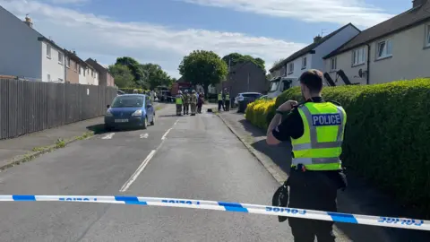 A uniformed police officer stands behind a cordon with more emergency service staff in the background
