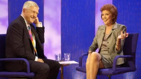 Sir Michael Parkinson pictured interviewing Cilla Black on his BBC One chat show in 2001. The pair sit opposite each other against a purple background