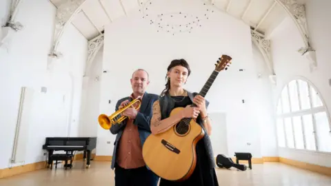 Guitarist Pippa Blundell - holding an acoustic dress and wearing a black dress - and trumpeter Colin Steele - in a dark jacket and pale orange shirt - standing inside a brightly lit building with white walls. 