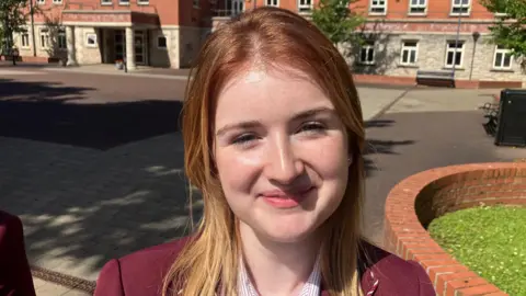 A young girl is smiling toward the camera. She has long blond hair. She is wearing a maroon coloured school blazer and a red and white stripe shirt. Behind her is an open stone courtyard and a number of red brick buildings.