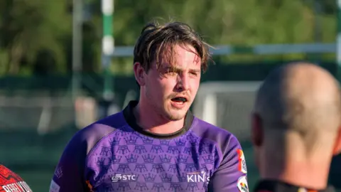 Colchester Kings Tom Crudgington during a rugby game. He is wearing a blue rugby shirt and a gumshield and his brown hair is ruffled. He has a short brown beard and a moustache. The back of another player's head is also in the shot.