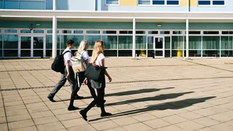 Three teenagers walk accross a paved area towards a school building wearing school uniform and rucksacks