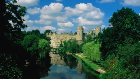 Getty Images A medieval castle with towers and turrets at the edge of a river, seen on a sunny day from a nearby bridge. Trees line the river bank