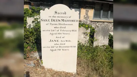 The Regency Society An old gravestone that has been freshly cleaned and restored. The sun is shining.