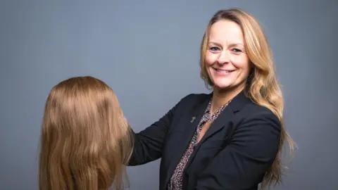 Wendy Tarplee-Morris is holding a light brown wig made from human hair by the charity she founded. Wendy is wearing a dark blue jacket over a brown, white and black blouse.