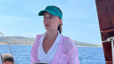 Family handout The image shows a teenage Tilly Seccombe on a yacht. She is wearing a turquoise sun visor on her head and a pink candy-striped blouse over a white halter-neck top.