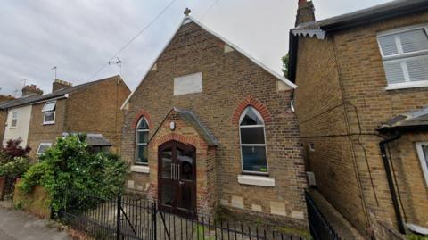 A Methodist church building. There is a small cross on the roof. The doors are brown wood and it has two lancet windows. The building sits in between terraced residential houses.