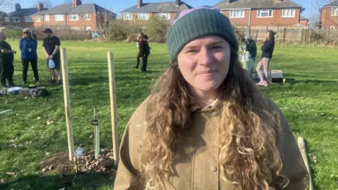 Woman with long brown curly hair wearing a purple and green beanie and a wax beige jacket. The tree stands behind her - with other people in the park in the background.