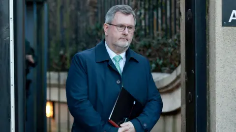 PA Media Jeffrey Donaldson exiting Newry courthouse carrying a notepad and phone. He is wearing a blue coat over a shirt and tie. He is looking off camera with a neutral expression. 