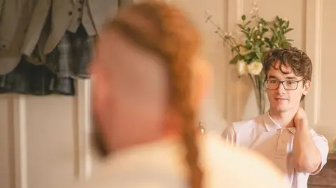 Duncan Ireland Photography A candid wedding photo. A young man is standing in his shirt with one arm around his own neck. In the foreground is the out-of-focus image of the rear of a large man with a pony tail. 