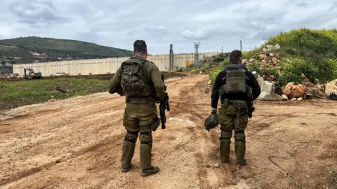 Reuters Two soldiers look at a concrete border wall at the end of a muddy track on a cloudy day.
