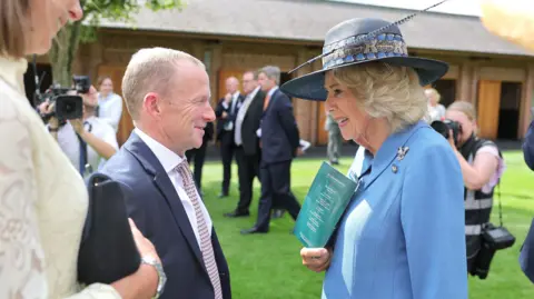 Several people are gathered outdoors on a lawn in front of a brick building during a formal event. In the foreground, two people are engaged in conversation. One person is wearing a light blue jacket and a wide-brimmed dark hat decorated with ribbon and a feather, and is holding a turquoise event programme. The other person is wearing a dark suit with a patterned tie. In the background, additional attendees in formal attire are standing and talking, while photographers and a camera operator capture the event. The scene is brightly lit by sunlight, with green grass and trees adding to the setting. Visible text on the programme is too small to read clearly.