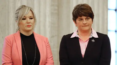 Michelle O'Neill standing next to Arlene Foster. O'Neill has blonde hair tied back with a fringe wearing a pink blazer and black top, and a long silver necklace. She is wearing makeup with pink lipstick. Foster has short brown hair and is wearing a pink striped shirt with a black blazer.