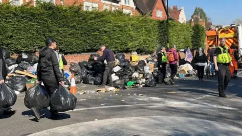 People carrying bin bags with a police officer in reflective vest watching and a bin lorry in the background. One man wearing a black jacket and a black bandana carries a bin bag in each hand as he walks towards a bin lorry. a man wearing a high-vis green jacket looks towards him.