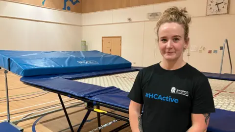 James Grant/BBC A woman with tied up blonde hair wearing a black t-shirt stands next to a wide trampoline with blue trim in a sports hall. 