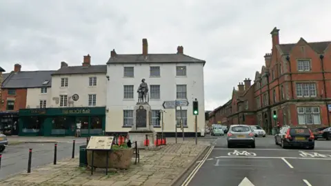 Google Streetview image of Alfreton market place, showing an open area with a war memorial, with a road and traditional shops running alongside