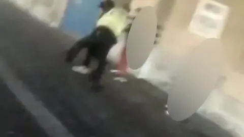 Image of a police officer in yellow arresting a teen people are blurred around the pair