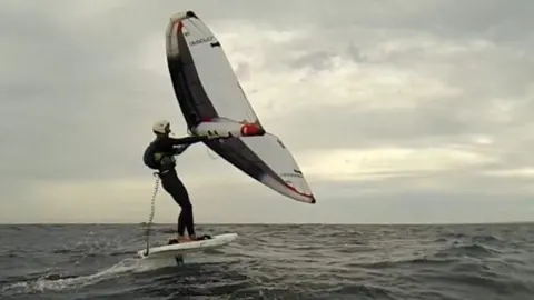 Martin Gaveriaux A wingfoiler stands balanced on a hydrofoil board above choppy sea, holding an inflated wing overhead while riding across the water under a cloudy sky.