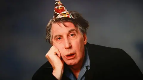 Scottish actor Rikki Fulton, poses with a characteristic glum expression. He has his face resting on his fist and is wearing a cone-shaped party hat on his head.