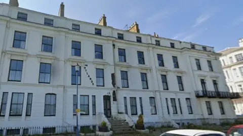 Google A large Victorian four-storey building with white walls and several windows with black frames.