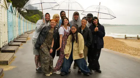 BBC/DSP/Georgina Vincent 10 people stood in a huddle with umbrellas on a beach front with beach huts to their left