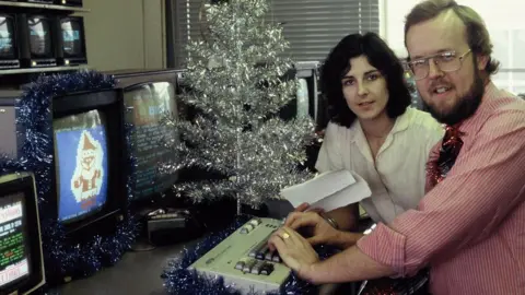 Liz Holdaway with dark shoulder-length hair and Ian Morton-Smith with a beard and glasses, at a desk with a Ceefax keyboard in front, several monitors, one showing a Ceefax Father Christmas design, and a silver Christmas tree on the desk