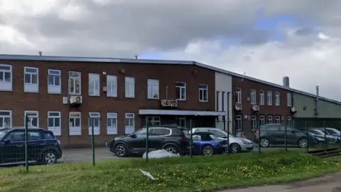 An outside view of the two storey Permoid Industries factory in Newton Aycliffe with a row of cars parked outside. The factory is made of brick with white windows. The roof rises to a central point at a shallow angle.
