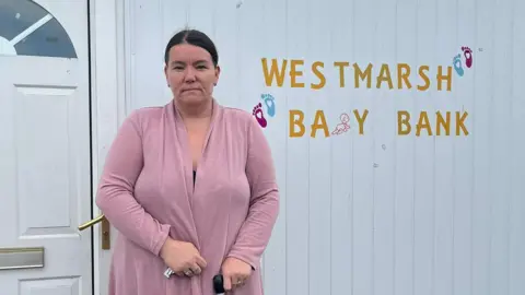 A woman with dark hair and wearing a pink jacket stands in front of a white cladded shipping container. There is gold coloured lettering on the wall which reads "West Marsh Baby Bank".