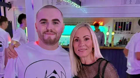 Family Handout Two people stand together in a bar or social venue with coloured lights overhead. One person is wearing a white T‑shirt featuring a large cartoon‑style character, while the other is dressed in a black, sparkly top. Other people and bar staff can be seen in the background.