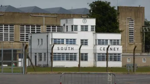 Creative Commons / Chris Allen The front of the Duke of York Barracks with a white building behind a wire fence with the words 'South Cerney' in front of a brown/grey building.