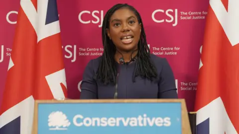 Kemi Badenoch gives a speech in front of a podium reading: "Conservatives". She is standing in front of a red backdrop reading: "CSJ The Centre for Social Justice" and two union jacks.
