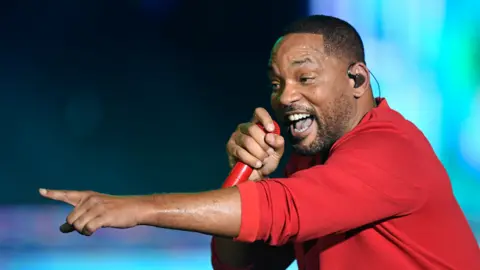 Will Smith is holding a microphone and wearing a red outfit on a stage, pointing to the left