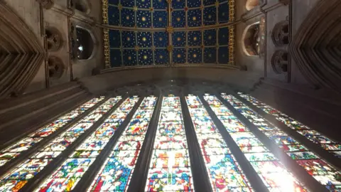 Looking up at Carlisle Cathedral's east window with the sun shining through, lighting up the array of colours. The decorated blue and gold starred ceiling can also be seen at the top of the picture.