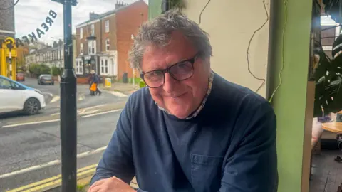 BBC/ Emily Johnson A man, who wears glasses and a navy jumper, smiles at a cafe table. He sits in a window seat which looks on to Bishopthorpe Road.